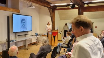 Die junge Frau am rednerpult. Auf dem Monitor wird nun ein gezeichnetes Portrait von einem Mann anzeigt.