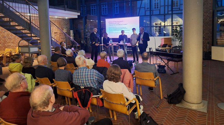 Eine Menschengruppe sitzen und von hinten fotografiert. Auf der Bühne fünf Personen stehend. Einer davon spricht in ein Mikrofon.