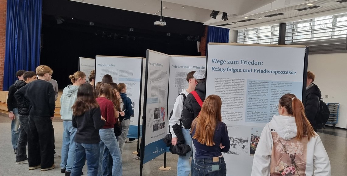 Schülerinnen und Schüler stehen in einem großen Raum vor Ausstellungswänden und betrachten diese.