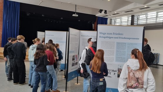 Schülerinnen und Schüler stehen in einem großen Raum vor Ausstellungswänden und betrachten diese.